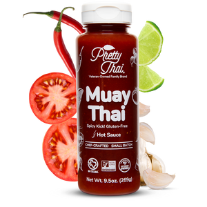 A bottle of Pretty Thai Muay Thai Stir Fry and dipping Sauce, which has savory with a thai chili kick and citrus tang. Net WT. 9.5 0z (269 g) is also written on it under different labels. In the background, different vegetables can be seen.