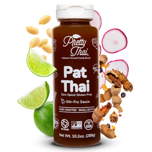 A bottle of Pretty Thai PAT Thai Stir Fry and Marinade Sauce, which hasIt has savory with a tangy punch of tamarind. Net WT. 10.2 0z (289 g) is also written on it under different labels. In the background, different nuts and vegetables can be seen.