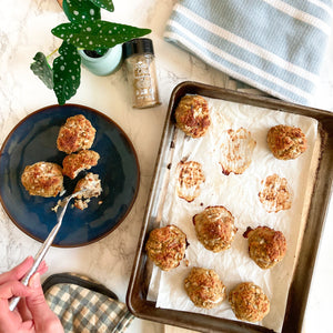 A tray with 7 meatballs. 3 meatballs are taken from the tray and put in the blue bowl. A human hand with a fork is on one of the meatballs. A white pot with plant in it, Pretty Thai’s seasoning salt bottle, a blue cloth with white stripes on it, and a grey and brown boxed cloth can also be seen.