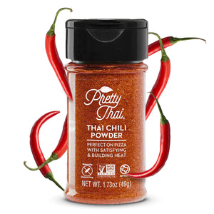 A bottle of Pretty Thai’s Thai Chili Powder. which will be perfect on pizza with satisfying and building heat. Net WT. 1.73 oz (49 g) is also written on it under different labels. In the background, red chilis can be seen.