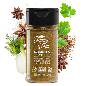 A bottle of Pretty Thai Seasoning Salt. which is for everything from the grill to the griddle. Net WT. 2 oz (57 g) is also written on it under different labels. In the background, different vegetables and organic products can be seen.
