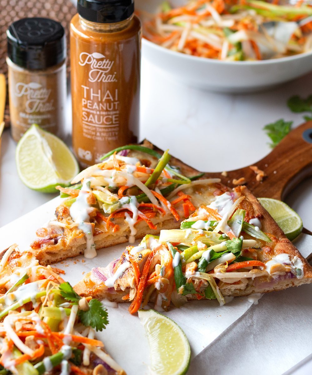 Two bottles of Pretty Thai products can be seen. One is small and the other one is Thai Peanut Dipping and Dressing Sauce. 3 slices of pizza are on the wooden board. Two pieces of lemon can also be seen.
