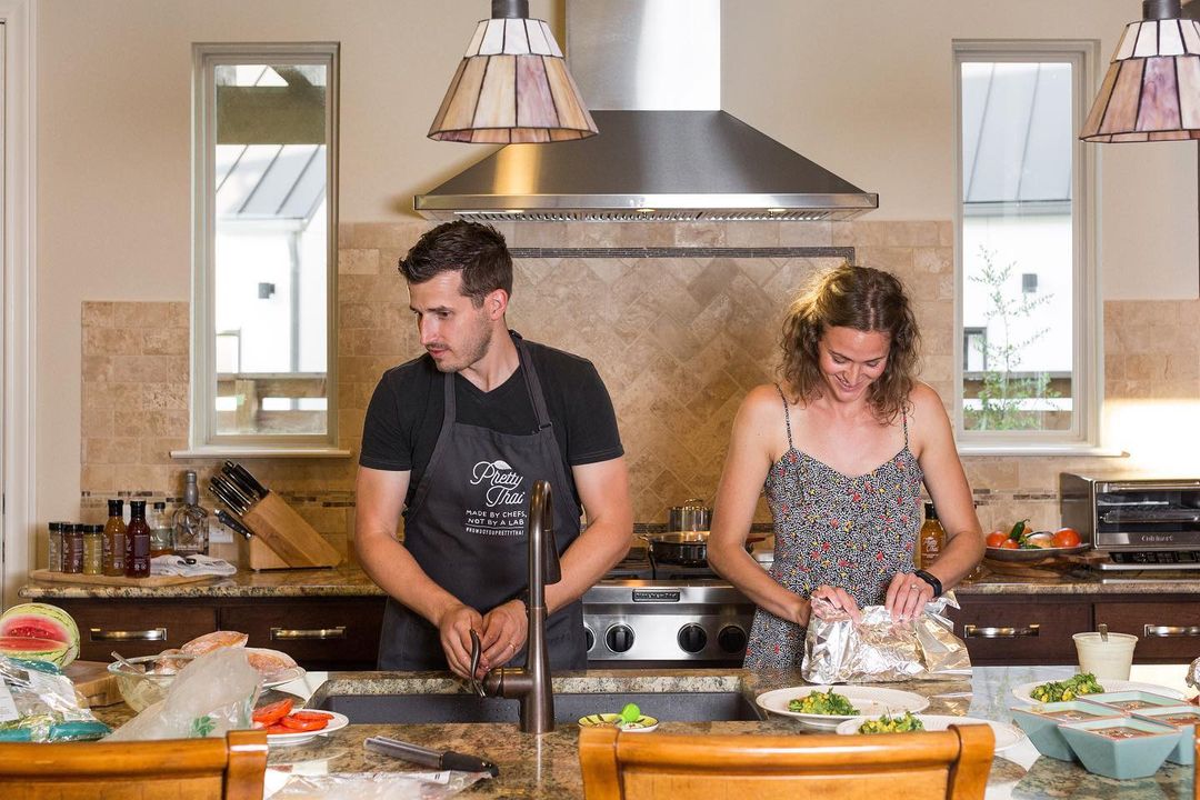 A Photo of couple in kitchen in which men is wearing Pretty Thai apran and women is opening some bag