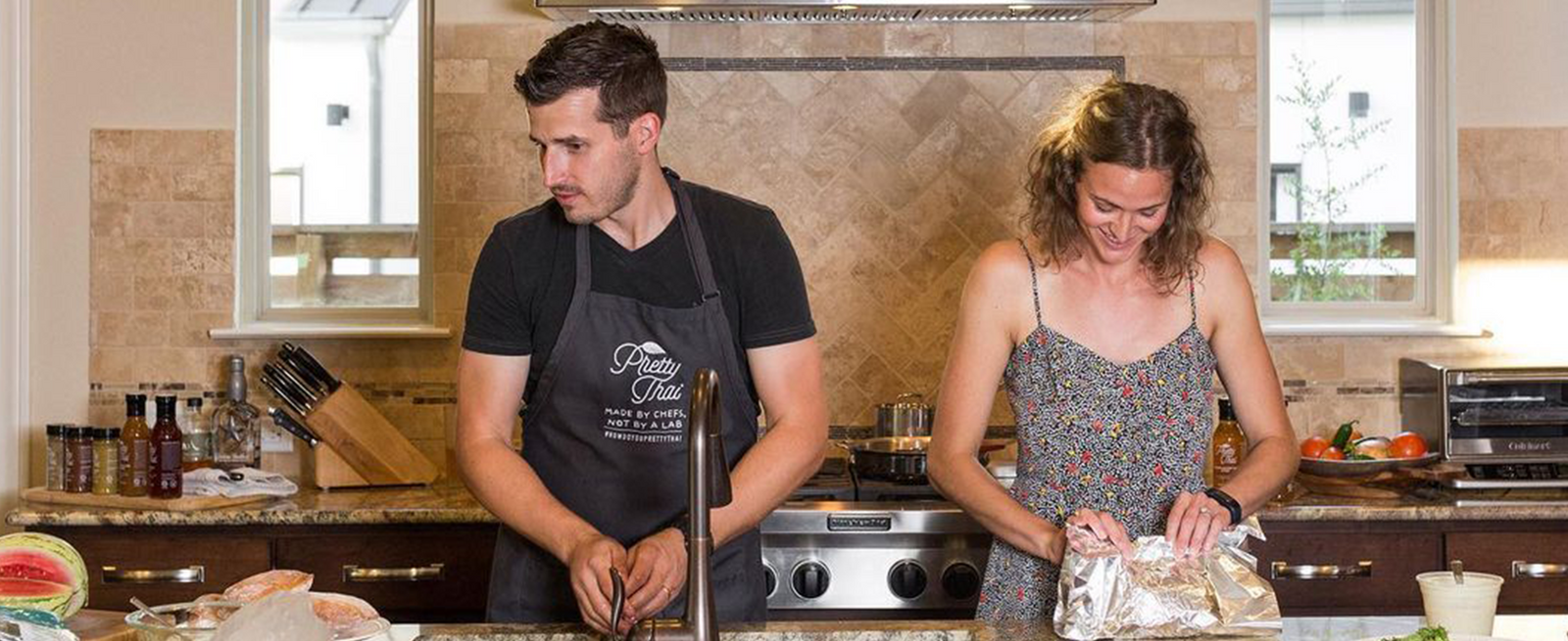 A Photo of couple in kitchen in which men is wearing Pretty Thai apran and women is opening some bag