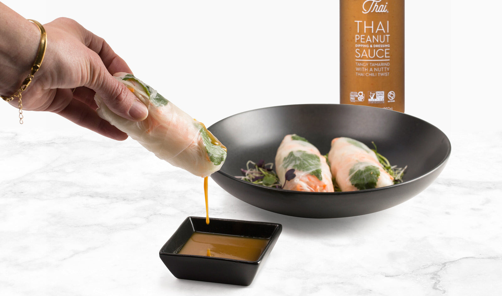 A black bowl with spring rolls on it and a women is dipping the roll in  thai peanut sauce