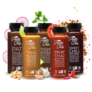  5 different bottles of Pretty Thai sauce flavors Including Pat Thai Sauce, Muay Thai Sauce, Thai Peanut Sauce, Sweet Chili Sauce, and Tiger Smirk Sauce.   In front of sweet chili stir fry and ba4rbecue sauce two red chili can be seen. Behind Muay Thai stir fry and dipping sauce a piece of tomato slice can be seen. In front of the tiger smirk grilling and dipping sauce, a clove of garlic, and behind the sauce mint leaves can be seen. In front of Pat thai stir fry and marinade sauce peanuts can be seen.