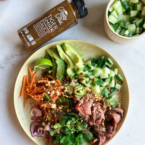 A big bowl with lemon, cucumber, onion, carrot, avocados, coriander, and other vegetables in it. A small bowl with pieces of cucumber in it. A bottle of Pretty Thai’s tiger smirk grilling and dripping sauce is lying with the big bowl. 