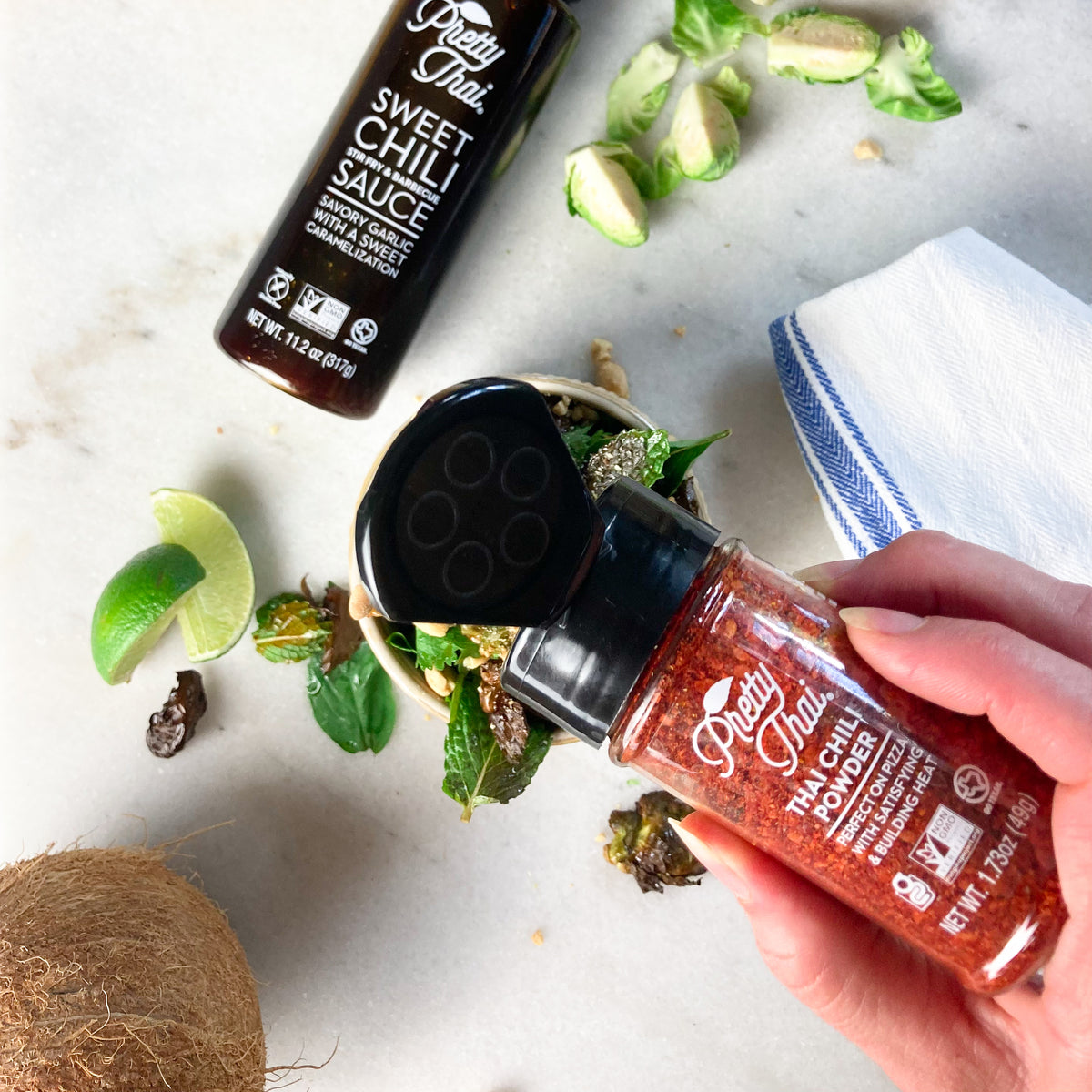 A white female hand is pouring Pretty Thai’s Thai Chili Powder. A bottle of sweet chili stir fry and barbecue sauce, a raw coconut, some lemon pieces, mint leaves, and a blue and white handkerchief can also be seen. 