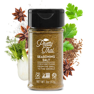 A bottle of Pretty Thai Seasoning Salt. which is for everything from the grill to the griddle. Net WT.  2 oz (57 g)  is also written on it under different labels. In the background, different vegetables and organic products can be seen.